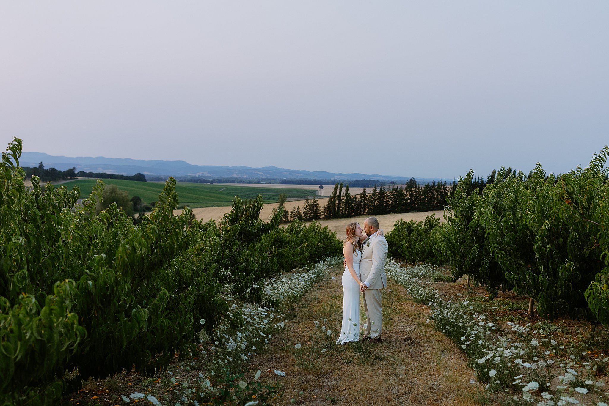 Bride and groom share an intimate kiss in a peaceful orchard at Perryhill Events, surrounded by lush green trees and rolling Oregon hills at sunset