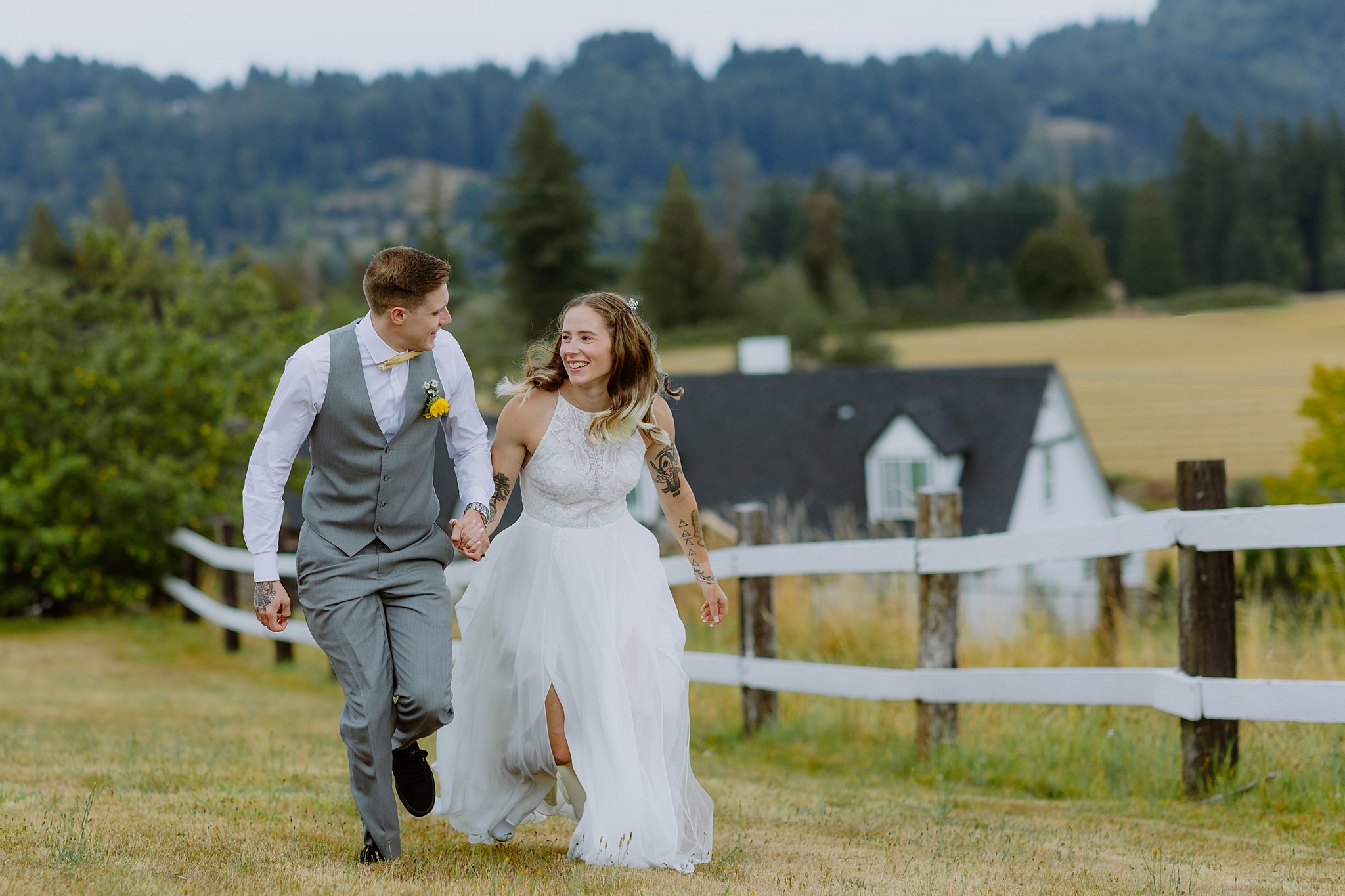 A joyful wedding couple is running hand-in-hand across a grassy field with a white fence in the foreground. The bride is wearing a flowing white halter-style dress with lace details, and the groom is dressed in a light gray vest and pants with a white shirt and yellow bowtie. Both are smiling and laughing, creating a lively, candid moment. In the background, a charming farmhouse and lush, rolling hills covered in trees complete the scenic countryside setting